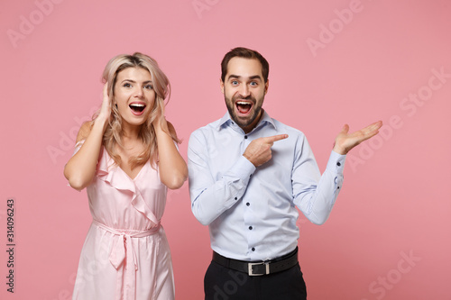 Excited young couple two guy girl in party outfit celebrating posing isolated on pastel pink background in studio. Valentine's Day Women's Day birthday holiday concept. Point index finger hand aside.