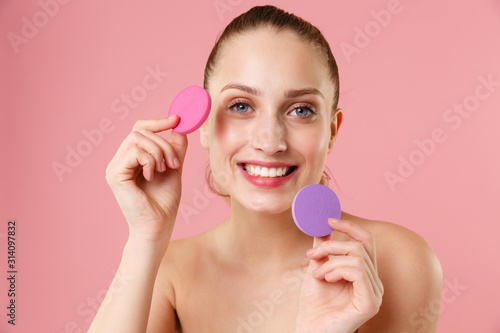 Close up blonde half naked woman 20s perfect skin blue eyes isolated on pastel pink wall background studio portrait. Skin care healthcare procedures concept Apply foundation nude makeup sponge on face