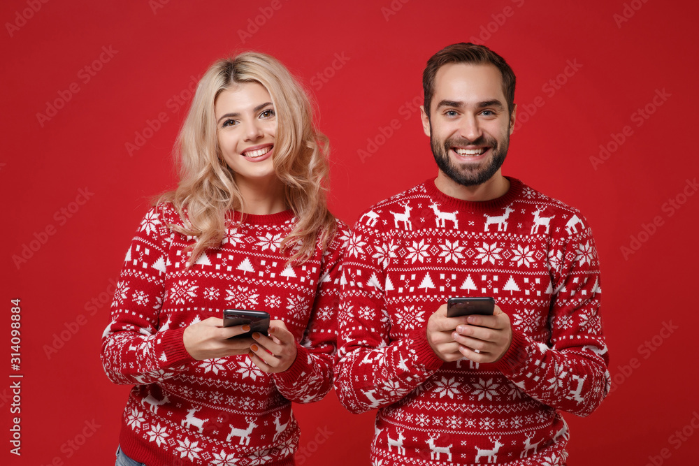 Beautiful young couple guy girl in Christmas knitted sweaters posing isolated on red background. New Year 2020 celebration holiday concept. Mock up copy space. Using mobile phone typing sms message.