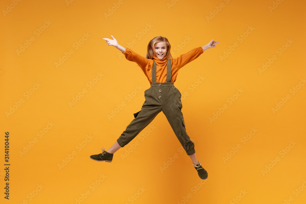 Smiling little kid girl 12-13 years old in turtleneck, jumpsuit isolated on orange yellow wall background in studio. Childhood lifestyle concept. Mock up copy space. Jumping, spreading hands and legs.