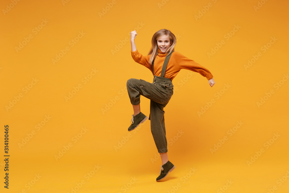 Joyful little blonde kid girl 12-13 years old in turtleneck, jumpsuit isolated on orange yellow background in studio. Childhood lifestyle concept. Mock up copy space. Rising leg, doing winner gesture.