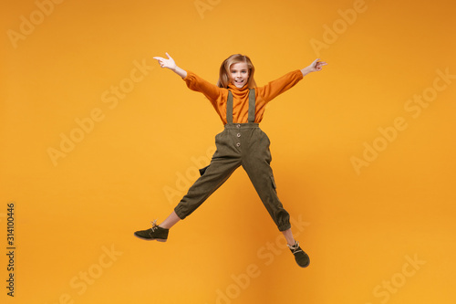 Smiling little kid girl 12-13 years old in turtleneck, jumpsuit isolated on orange yellow wall background in studio. Childhood lifestyle concept. Mock up copy space. Jumping, spreading hands and legs. © ViDi Studio
