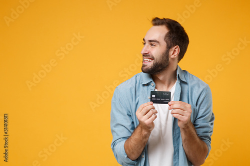 Smiling young man in casual blue shirt posing isolated on yellow orange background, studio portrait. People emotions lifestyle concept. Mock up copy space. Holding credit bank card, looking aside.