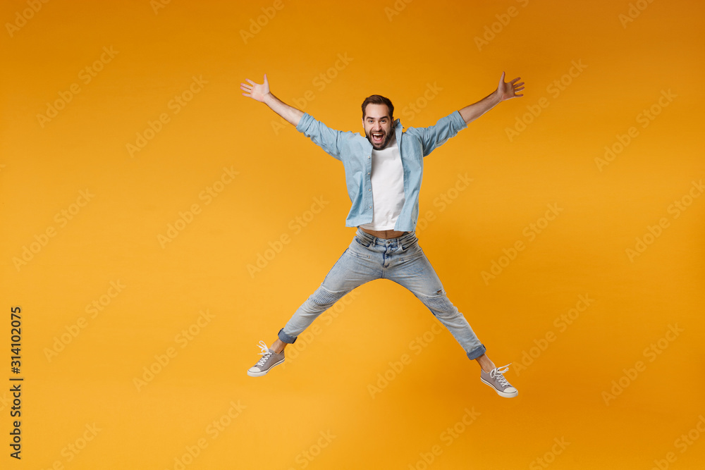 Joyful young bearded man in casual blue shirt posing isolated on yellow orange wall background studio portrait. People emotions lifestyle concept. Mock up copy space. Jumping, spreading hands, legs.