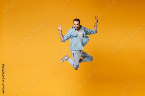 Smiling young bearded man in casual blue shirt posing isolated on yellow orange background studio portrait. People sincere emotions lifestyle concept. Mock up copy space. Jumping showing victory sign.