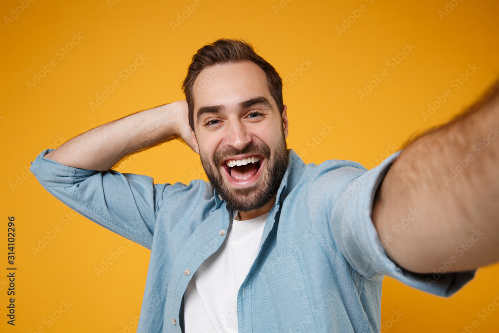 Close up of handsome funny young man in casual blue shirt posing isolated on yellow orange background, studio portrait. People lifestyle concept. Mock up copy space. Doing selfie shot on mobile phone. Close up of handsome funny young man in casual blue shirt posing isolated on yellow orange background, studio portrait. People lifestyle concept. Mock up copy space. Doing selfie shot on mobile phone.
