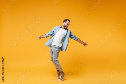 Laughing young bearded man in casual blue shirt posing isolated on yellow orange background studio portrait. People emotions lifestyle concept. Mock up copy space. Standing on toes, spreading hands. photo