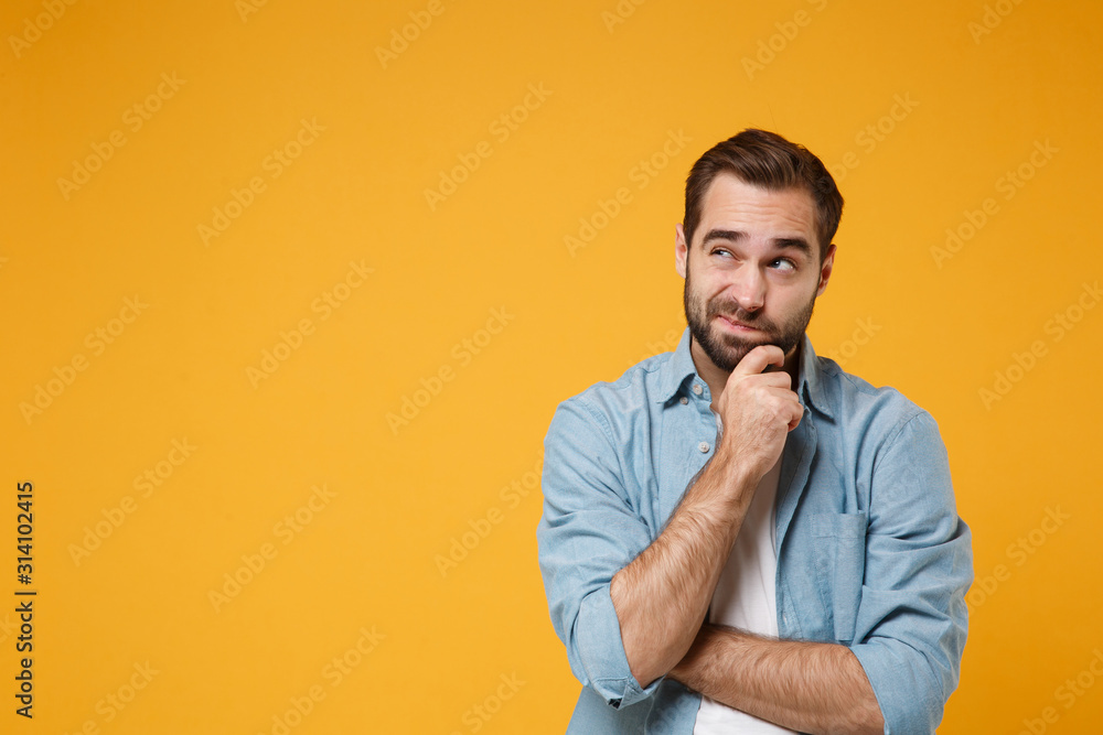 Pensive young bearded man in casual blue shirt posing isolated on yellow orange background studio portrait. People emotions lifestyle concept. Mock up copy space. Put hand prop up on chin, looking up. - obrazy, fototapety, plakaty 