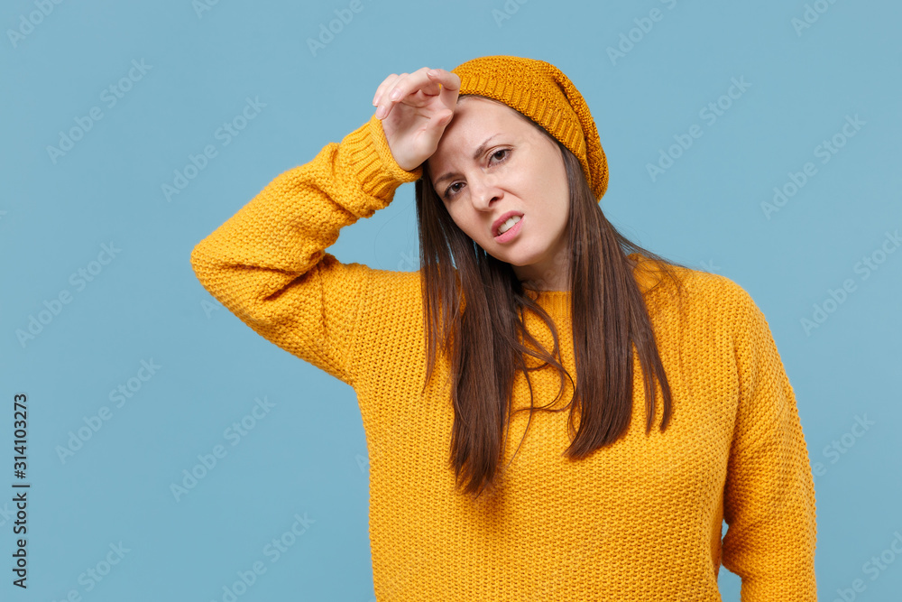 Exhausted tired young brunette woman girl in yellow sweater and hat posing isolated on blue wall background studio portrait. People emotions lifestyle concept. Mock up copy space. Put hand on head.
