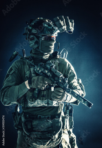 Anti-terrorist squad fighter, army elite forces soldier in combat uniform and tactical ammunition, armed mini submachine gun, wearing night-vision device, low key studio portrait on black background © Getmilitaryphotos