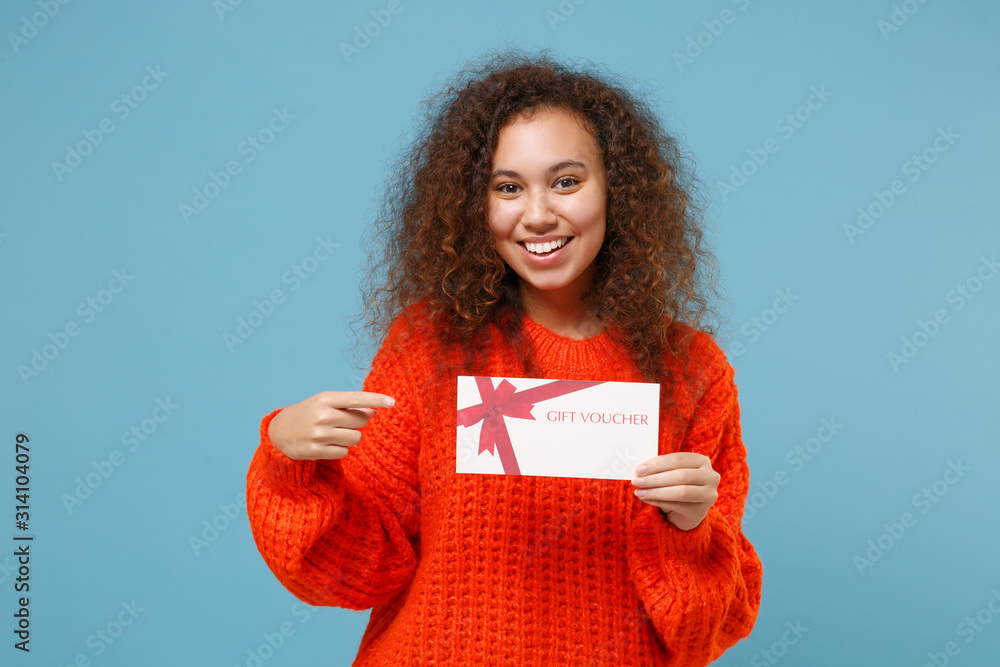 Pretty young african american girl in casual orange knitted clothes isolated on pastel blue wall background. People lifestyle concept. Mock up copy space. Pointing index finger on gift certificate. Pretty young african american girl in casual orange knitted clothes isolated on pastel blue wall background. People lifestyle concept. Mock up copy space. Pointing index finger on gift certificate.