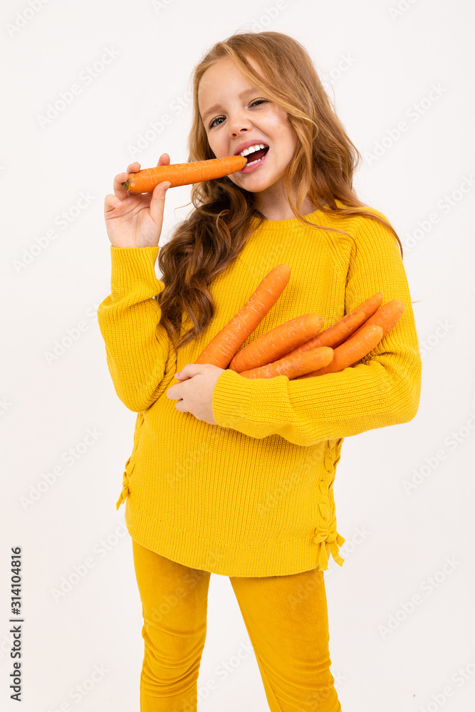 Happy teenager girl with red hair, hoody and yellow trousers eats carrot isolated on white background Happy teenager girl with red hair, hoody and yellow trousers eats carrot isolated on white background