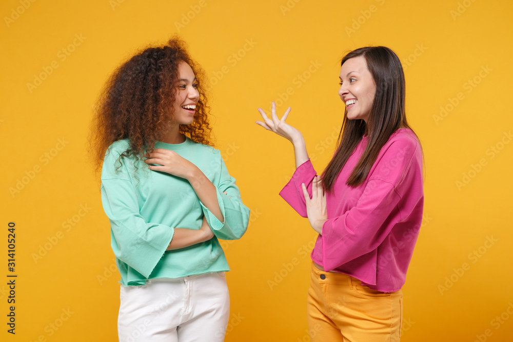 Two cheerful women friends european and african american girls in pink green clothes posing isolated on yellow background. People lifestyle concept. Mock up copy space. Looking at each other speaking. Two cheerful women friends european and african american girls in pink green clothes posing isolated on yellow background. People lifestyle concept. Mock up copy space. Looking at each other speaking.