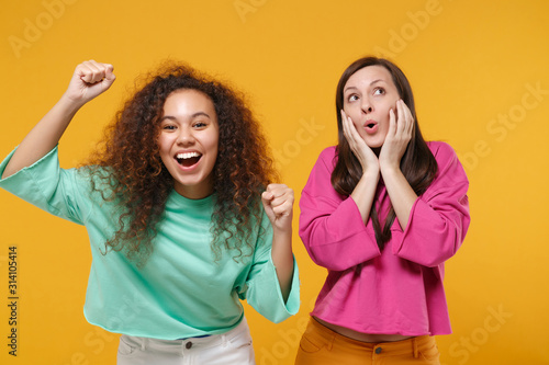 Two funny women friends european african american girls in pink green clothes posing isolated on yellow background. People lifestyle concept. Mock up copy space. Clenching fists put hands on cheeks.