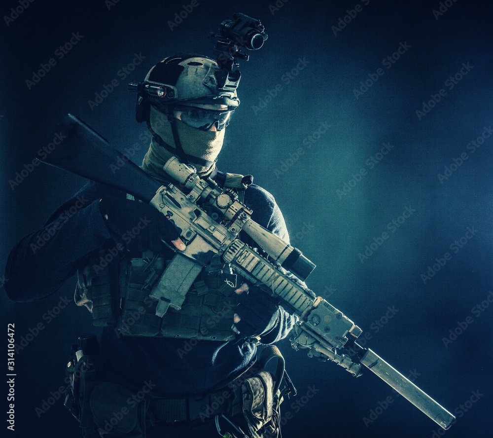 Naklejka premium Army special operations soldier, counter-terrorist crew sniper, marksman in combat helmet, hiding face behind mask, armored service rifle with optical sight, low key studio shoot on blue background