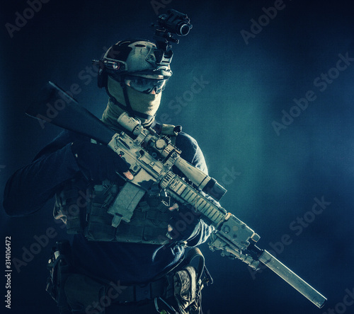 Army special operations soldier, counter-terrorist crew sniper, marksman in combat helmet, hiding face behind mask, armored service rifle with optical sight, low key studio shoot on blue background