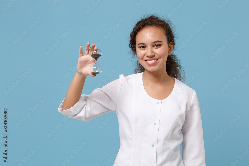 Smiling african american doctor woman isolated on blue wall background. Female doctor in white medical gown hold hourglass black sand. Healthcare personnel medicine health concept. Mock up copy space.