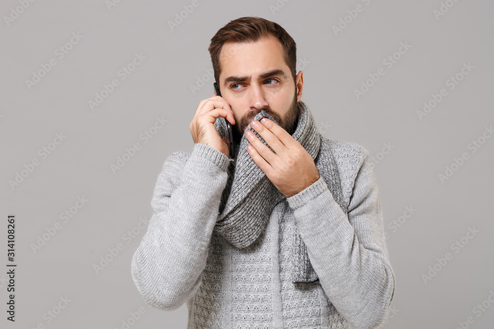 Man in gray sweater isolated on grey background. Healthy lifestyle ill sick disease treatment cold season concept. Mock up copy space. Talking on mobile phone coughing sneezing cover mouth with scarf. Man in gray sweater isolated on grey background. Healthy lifestyle ill sick disease treatment cold season concept. Mock up copy space. Talking on mobile phone coughing sneezing cover mouth with scarf.