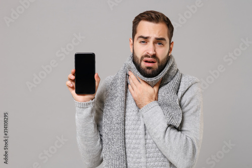 Man in gray sweater, scarf isolated on grey background. Healthy lifestyle, ill sick disease treatment, cold season concept. Mock up copy space. Hold mobile phone with blank screen, put hand on throat.