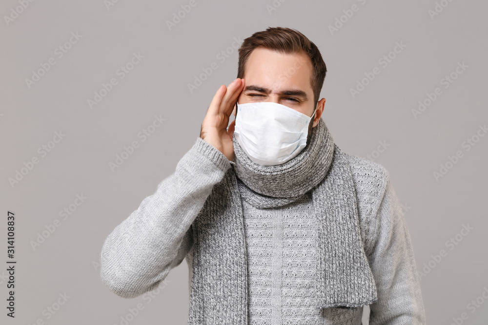 Young man in gray sweater, scarf isolated on grey background. Healthy lifestyle, ill sick disease treatment, cold season concept. Mock up copy space. Wearing sterile face mask put hand on temple head. Young man in gray sweater, scarf isolated on grey background. Healthy lifestyle, ill sick disease treatment, cold season concept. Mock up copy space. Wearing sterile face mask put hand on temple head.