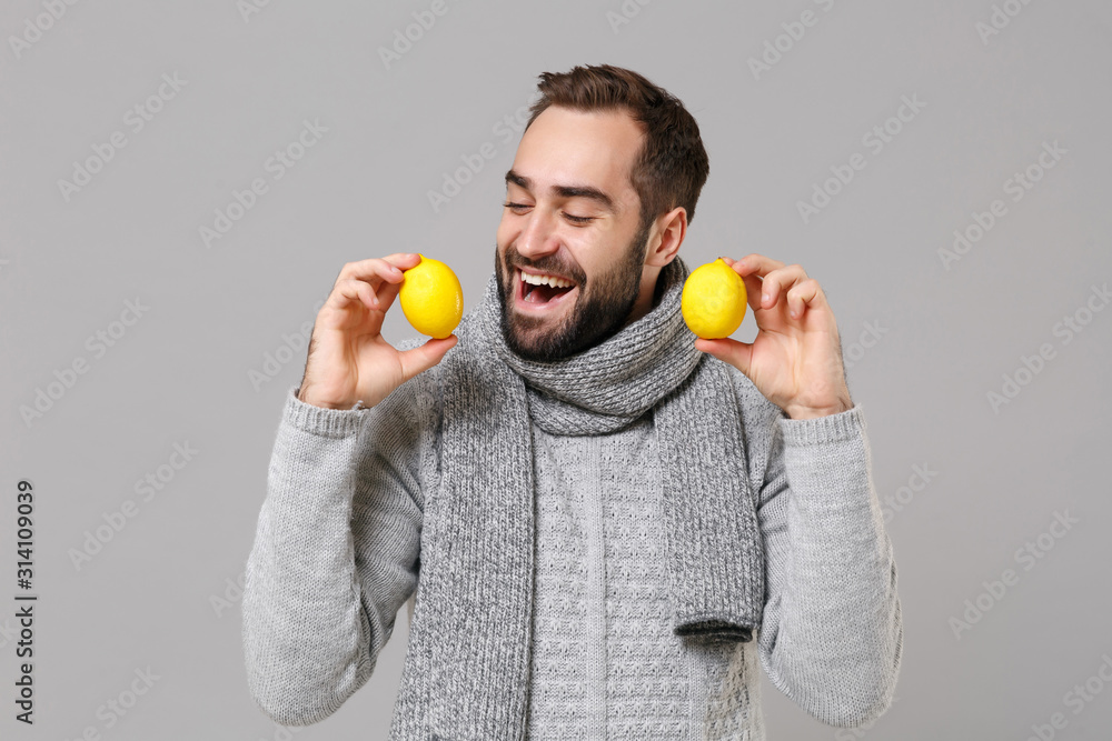 Cheerful young man in gray sweater, scarf posing isolated on grey wall background, studio portrait. Healthy fashion lifestyle, people emotions, cold season concept. Mock up copy space. Holding lemons. Cheerful young man in gray sweater, scarf posing isolated on grey wall background, studio portrait. Healthy fashion lifestyle, people emotions, cold season concept. Mock up copy space. Holding lemons.