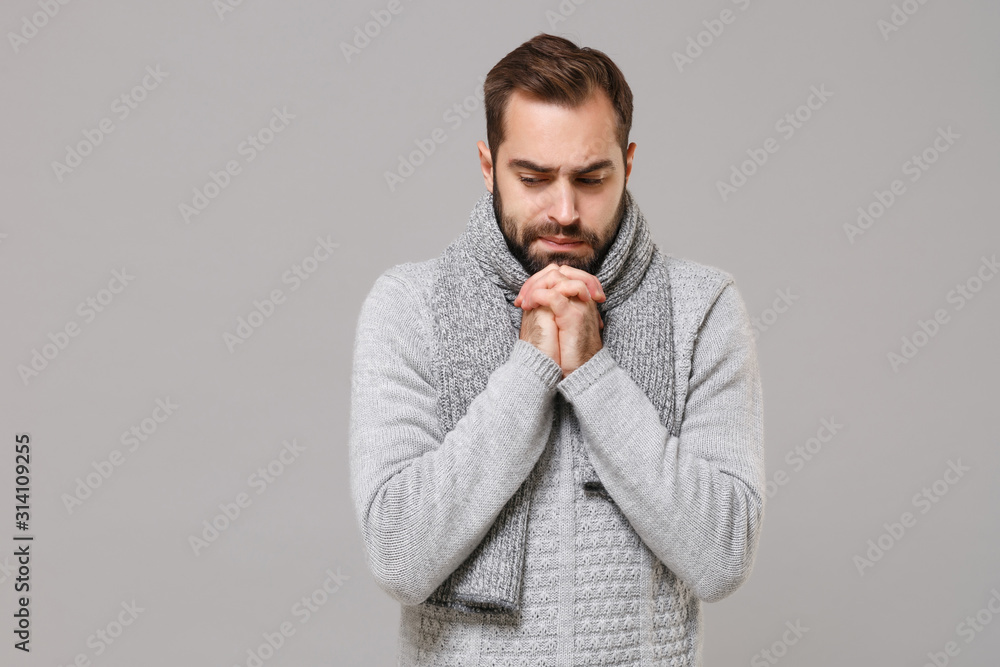 Young man in gray sweater, scarf posing isolated on grey background, studio portrait. Healthy fashion lifestyle, people emotions, cold season concept. Mock up copy space. Hold hands folded in prayer. Young man in gray sweater, scarf posing isolated on grey background, studio portrait. Healthy fashion lifestyle, people emotions, cold season concept. Mock up copy space. Hold hands folded in prayer.