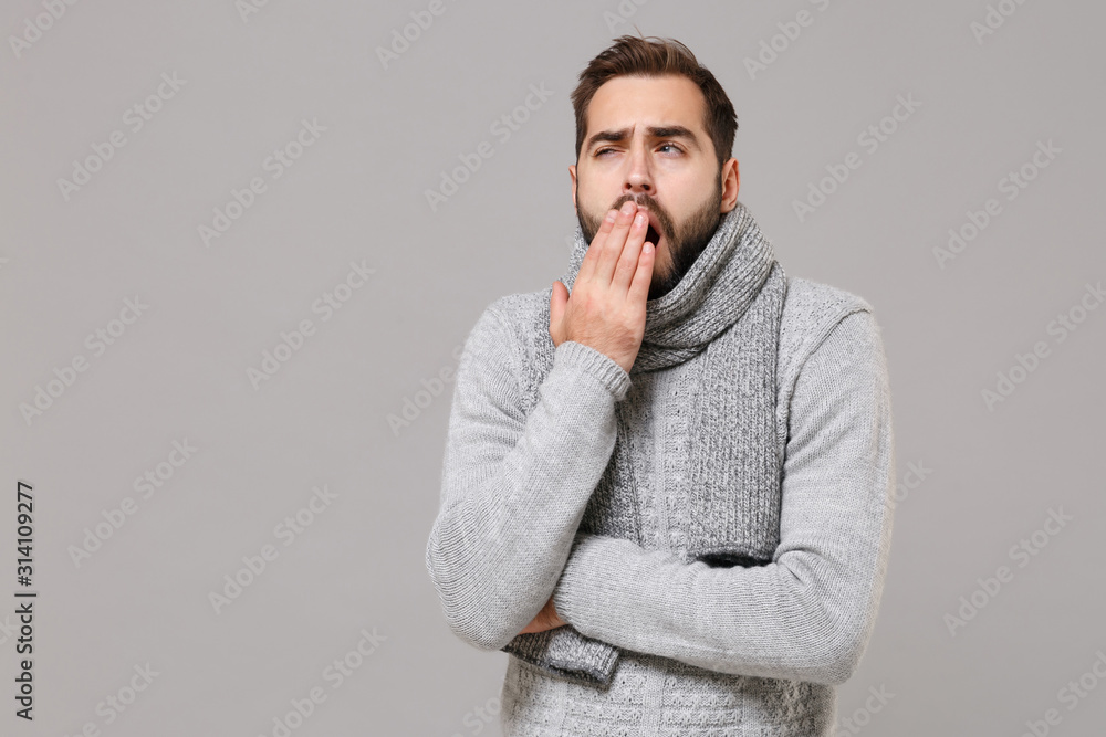 Tired young man in gray sweater, scarf posing isolated on grey wall background, studio portrait. Healthy fashion lifestyle, cold season concept. Mock up copy space. Yawning, covering mouth with hand. Tired young man in gray sweater, scarf posing isolated on grey wall background, studio portrait. Healthy fashion lifestyle, cold season concept. Mock up copy space. Yawning, covering mouth with hand.