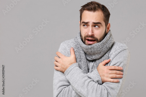 Frozen young man in gray sweater posing isolated on grey wall background, studio portrait. Healthy fashion lifestyle, cold season concept. Mock up copy space. Holding hands folded, huging himself. photo