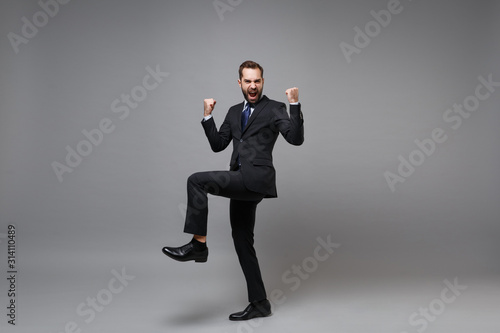 Overjoyed young business man in classic black suit shirt tie posing isolated on grey background. Achievement career wealth business concept. Mock up copy space. Clenching fists like winner, screaming.