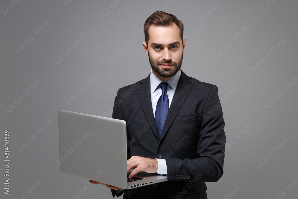 Confident young business man in classic black suit shirt tie posing isolated on grey background. Achievement career wealth business concept. Mock up copy space. Holding, working on laptop pc computer. Confident young business man in classic black suit shirt tie posing isolated on grey background. Achievement career wealth business concept. Mock up copy space. Holding, working on laptop pc computer.