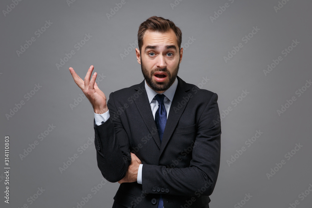 Perplexed irritated young bearded business man in classic black suit shirt tie posing isolated on grey wall background. Achievement career wealth business concept. Mock up copy space. Spreading hands. Perplexed irritated young bearded business man in classic black suit shirt tie posing isolated on grey wall background. Achievement career wealth business concept. Mock up copy space. Spreading hands.