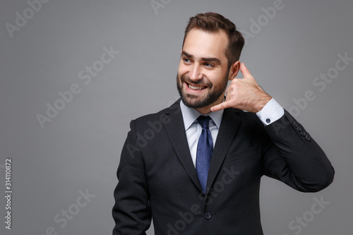 Smiling young business man in classic suit shirt tie posing isolated on grey background. Achievement career wealth business concept. Mock up copy space. Doing phone gesture like says call me back.