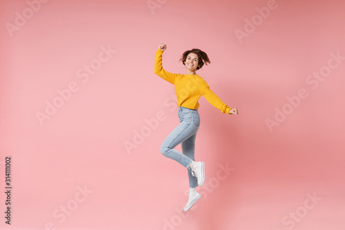 Joyful young brunette woman girl in yellow sweater posing isolated on pastel pink wall background. People lifestyle concept. Mock up copy space. Having fun fooling around jumping doing winner gesture.