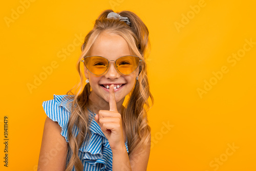 charming blonde girl in a dress and sunglasses asks to be quieter, holds a finger at her mouth on a yellow background with copy space