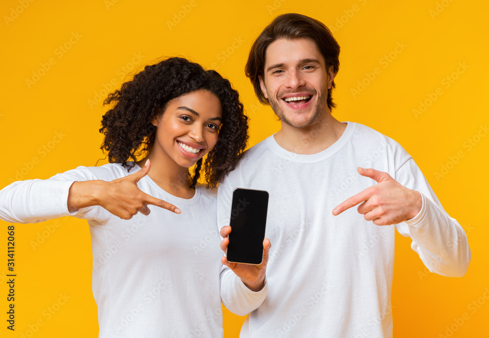 Cheerful interracial couple pointing at smartphone with black screen for mockup - obrazy, fototapety, plakaty 