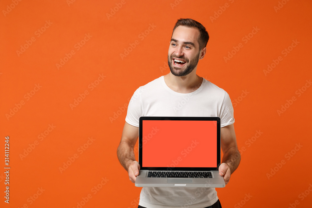 Cheerful young man in casual white t-shirt posing isolated on bright orange background studio portrait. People lifestyle concept. Mock up copy space. Hold laptop pc computer with blank empty screen. Cheerful young man in casual white t-shirt posing isolated on bright orange background studio portrait. People lifestyle concept. Mock up copy space. Hold laptop pc computer with blank empty screen.