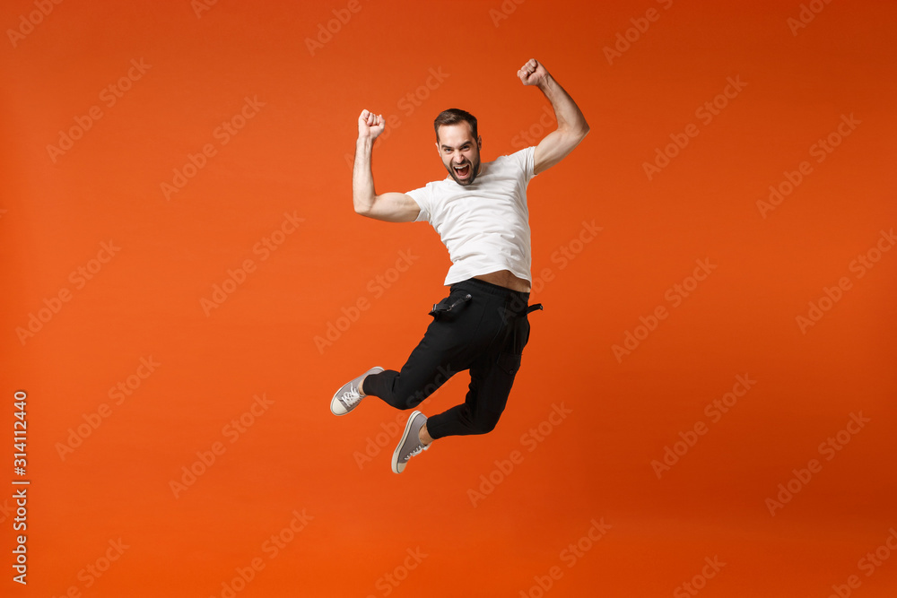 Crazy young man in casual white t-shirt posing isolated on orange background in studio. People lifestyle concept. Mock up copy space. Having fun, fooling around, jumping, clenching fists like winner.
