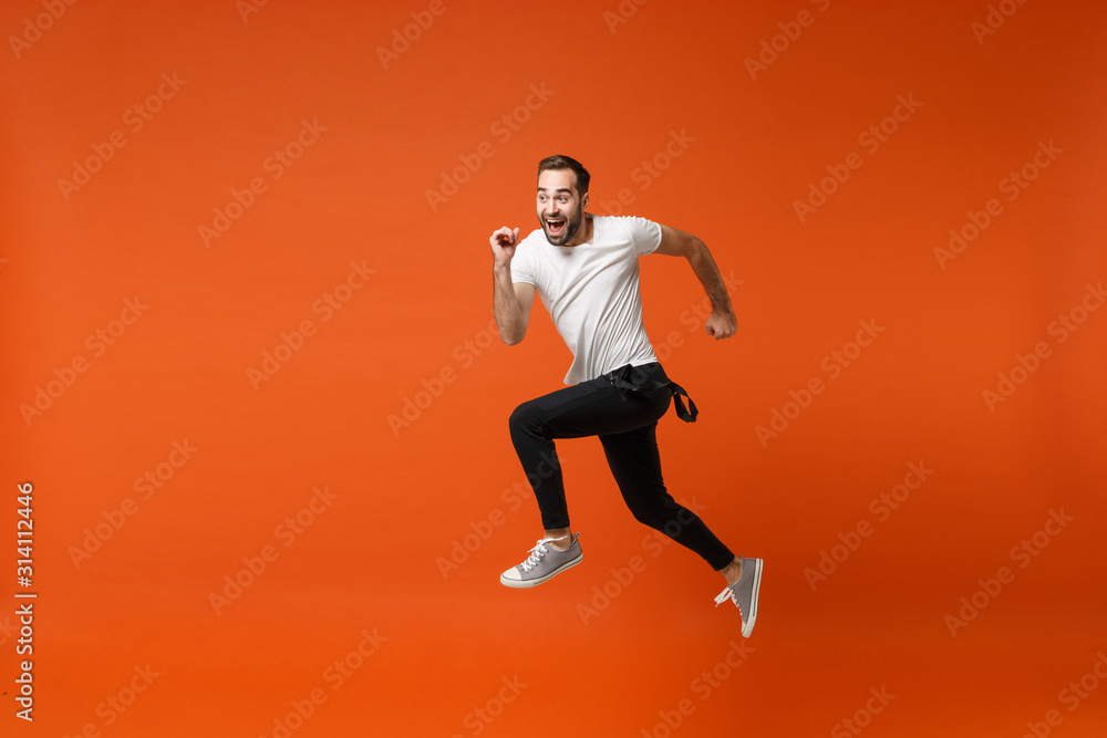 Cheerful funny young man in casual white t-shirt posing isolated on bright orange wall background studio portrait. People lifestyle concept. Mock up copy space. Having fun, fooling around, jumping.