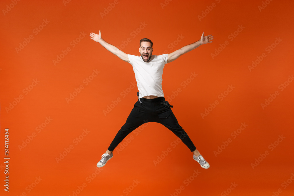 Joyful young man in casual white t-shirt posing isolated on orange background studio portrait. People sincere emotions lifestyle concept. Mock up copy space. Have fun jumping spreading hands and legs.
