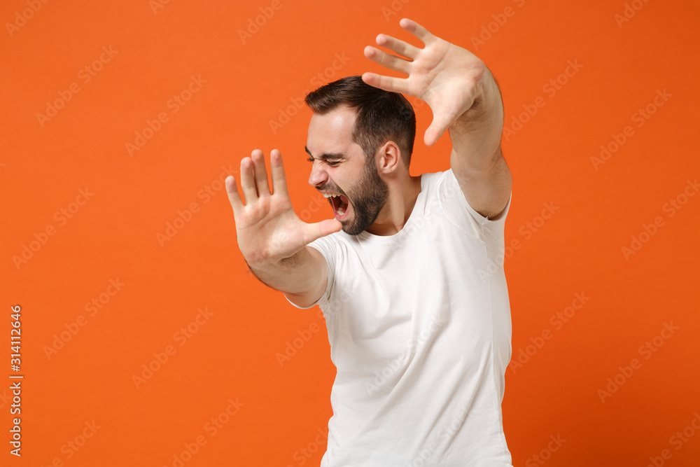 Scared displeased young man in casual white t-shirt posing isolated on orange background, studio portrait. People sincere emotions lifestyle concept. Mock up copy space. Screaming covering with hands. Scared displeased young man in casual white t-shirt posing isolated on orange background, studio portrait. People sincere emotions lifestyle concept. Mock up copy space. Screaming covering with hands.