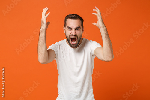 Angry young man in casual white t-shirt posing isolated on bright orange wall background studio portrait. People sincere emotions lifestyle concept. Mock up copy space. Screaming, spreading hands.