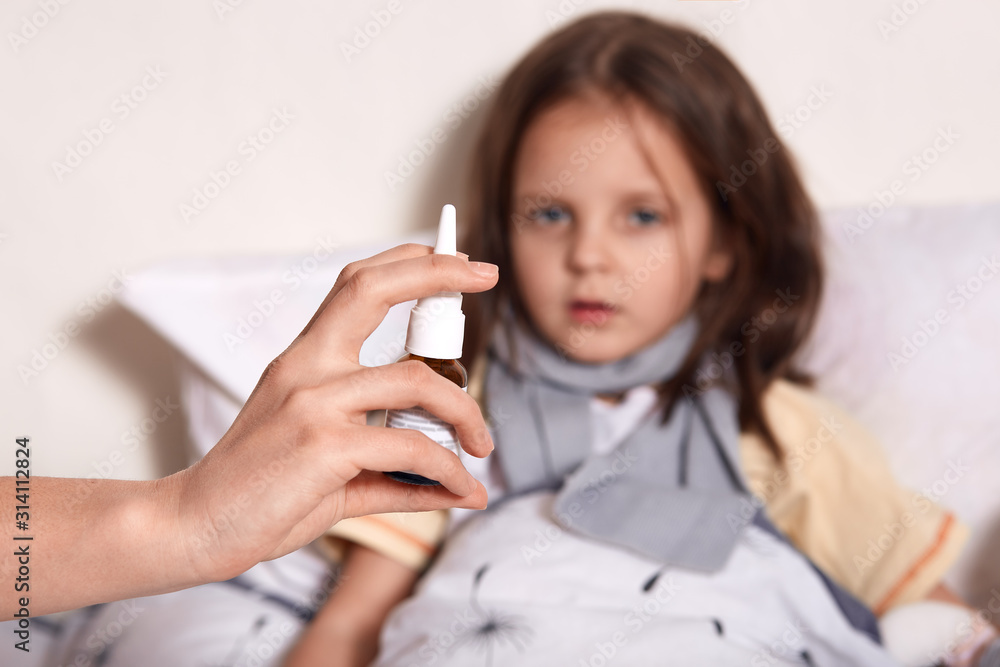 Horizontal shot of little girl lying in bed, her mom treting her ranning nose with nasal spray, dark haired female child looking at camera, looks ill, suffers from flue. Health care concept. Horizontal shot of little girl lying in bed, her mom treting her ranning nose with nasal spray, dark haired female child looking at camera, looks ill, suffers from flue. Health care concept.