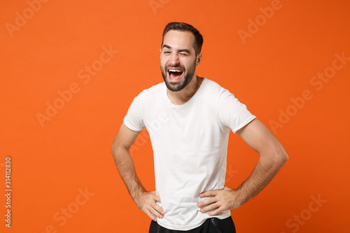 Funny young man in casual white t-shirt posing isolated on orange wall background, studio portrait. People sincere emotions lifestyle concept. Mock up copy space. Standing with arms akimbo, blinking.