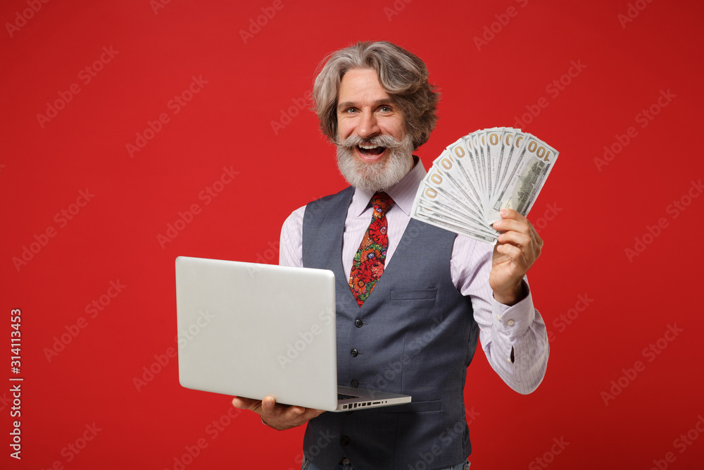 Smiling elderly gray-haired mustache bearded man in classic shirt vest tie posing isolated on red background. People lifestyle concept. Mock up copy space. Hold laptop pc computer, fan of cash money. Smiling elderly gray-haired mustache bearded man in classic shirt vest tie posing isolated on red background. People lifestyle concept. Mock up copy space. Hold laptop pc computer, fan of cash money.