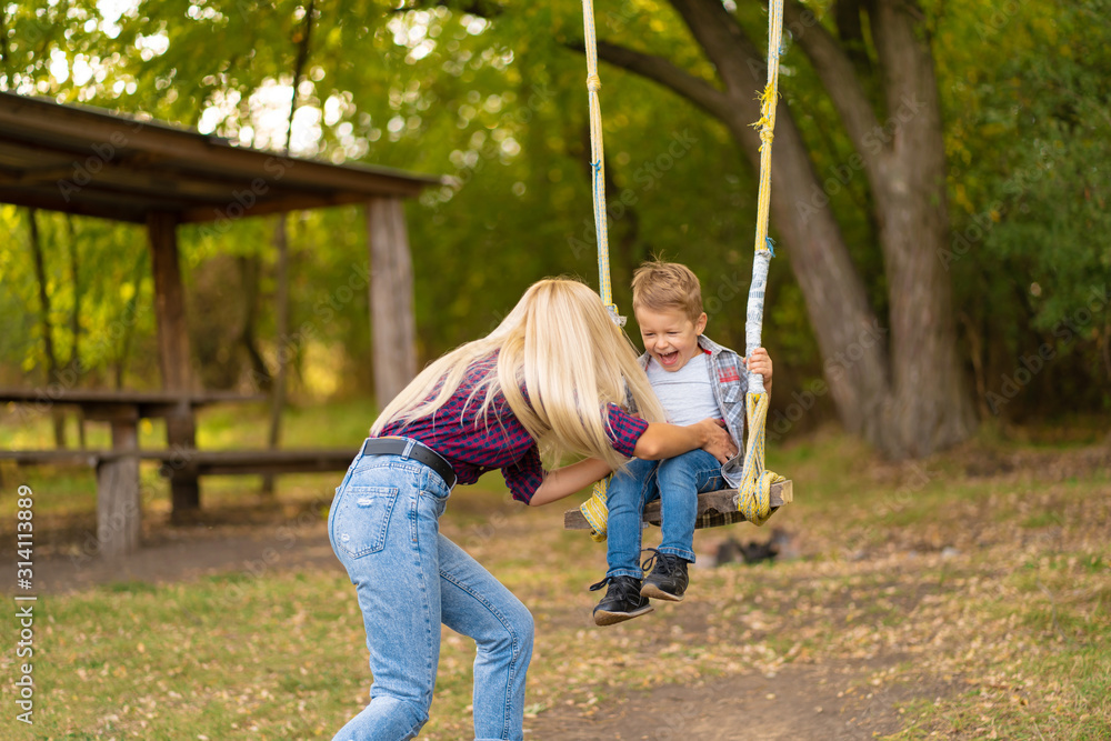 Young blonde mom shakes her little son on a swing in a green park. Happy childhood.