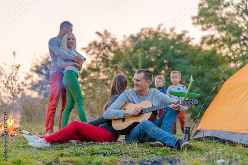 Group of happy friends with guitar, having fun outdoor, near bonfire and tourist tent. Camping fun happy family