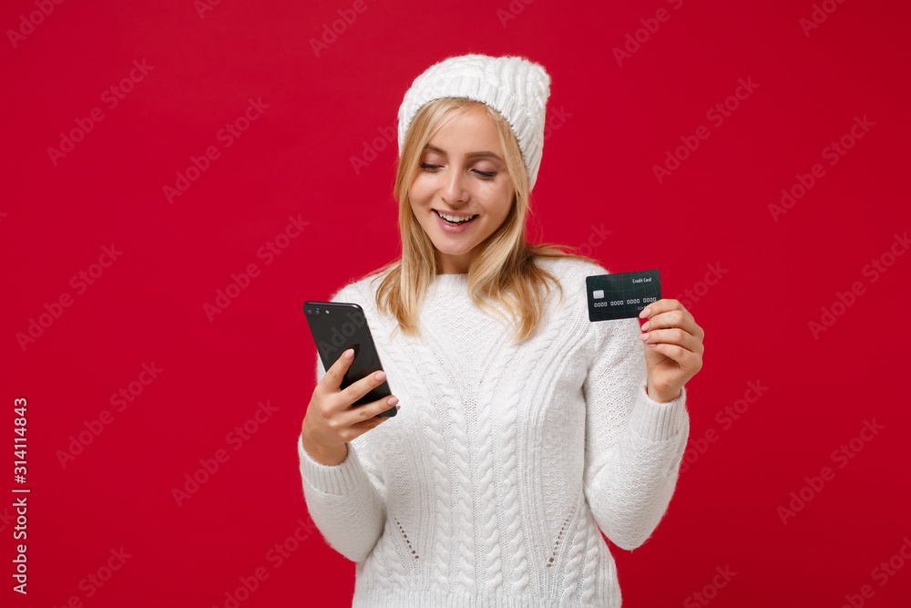 Pretty young woman in white sweater, hat isolated on red wall background, studio portrait. Healthy fashion lifestyle, cold season concept. Mock up copy space. Holding mobile phone, credit bank card. Pretty young woman in white sweater, hat isolated on red wall background, studio portrait. Healthy fashion lifestyle, cold season concept. Mock up copy space. Holding mobile phone, credit bank card.