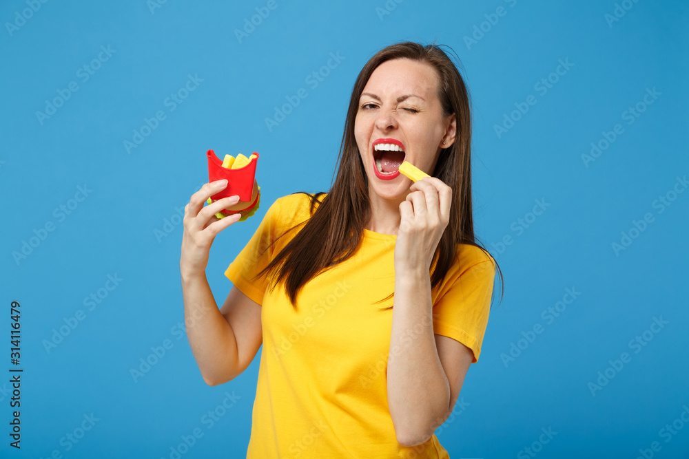 Funny young brunette woman girl in yellow t-shirt posing isolated on blue background, studio portrait. People lifestyle concept. Mock up copy space. Hold plastic toys french fries potatoes burger. Funny young brunette woman girl in yellow t-shirt posing isolated on blue background, studio portrait. People lifestyle concept. Mock up copy space. Hold plastic toys french fries potatoes burger.