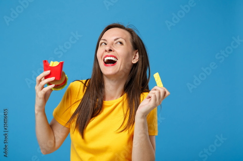 Laughing young brunette woman girl in yellow t-shirt posing isolated on blue background, studio portrait. People lifestyle concept. Mock up copy space. Hold plastic toys french fries potatoes burger. photo