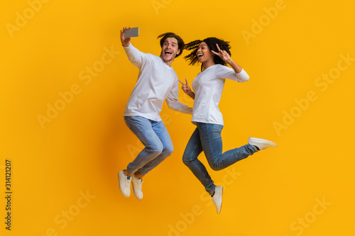 Millennial Interracial Couple Jumping And Taking Selfiem Posing Over Yellow Background